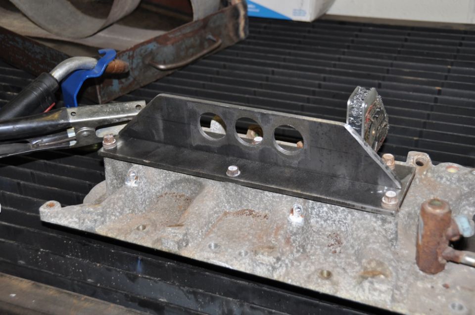 Custom metal bracket with three holes bolted onto an aluminum intake manifold
