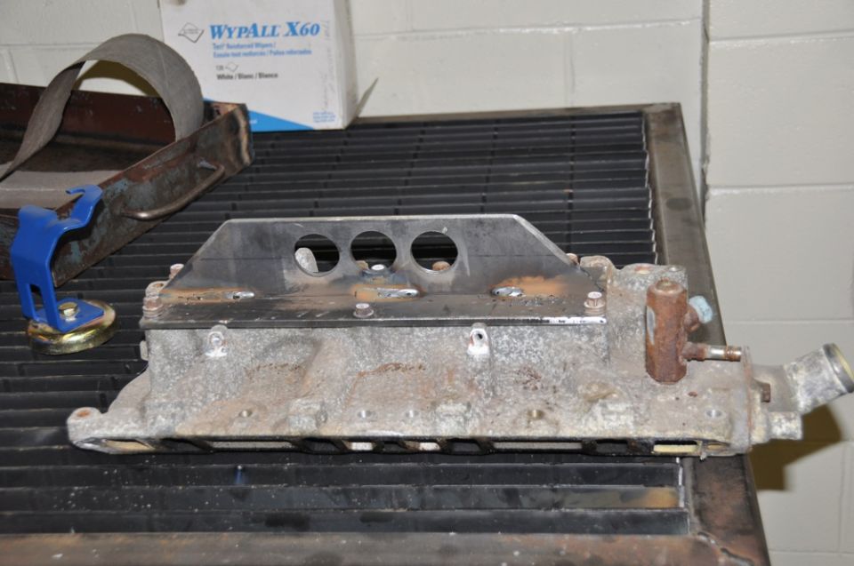 Engine intake manifold with a metal bracket bolted on top, sitting on a black grate surface. A box of WypAll X60 wipes is in the background