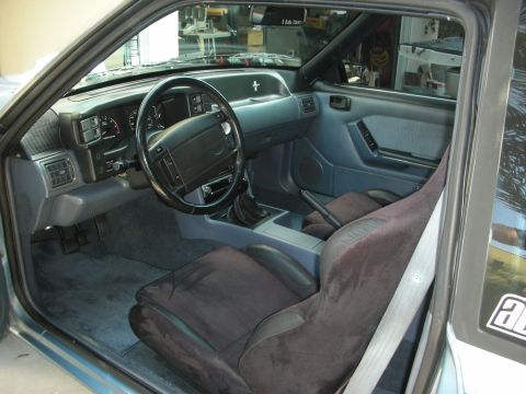 1990 Ford Mustang - The interior of a 1990 Ford Mustang shows a grey dashboard, black steering wheel, and dark fabric seats.
