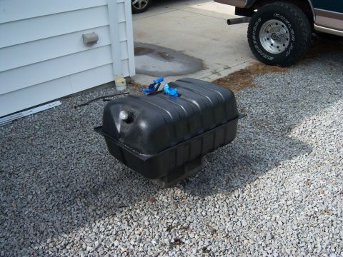 1996 Ford Bronco - A black plastic fuel tank with blue fittings sits on a gravel surface next to a white sided building.