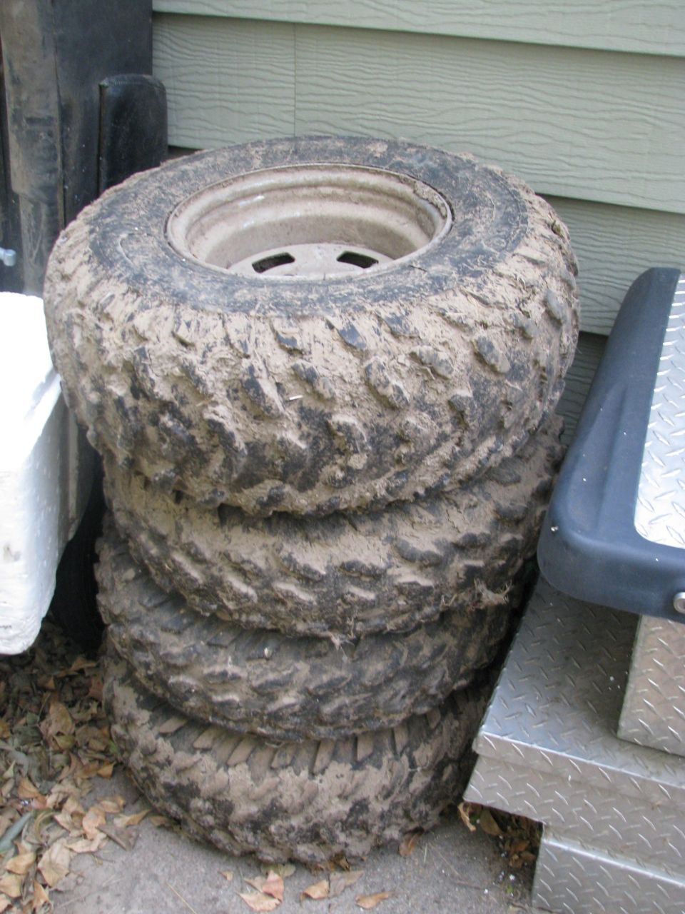 Four muddy off-road tires on white steel wheels stacked outdoors