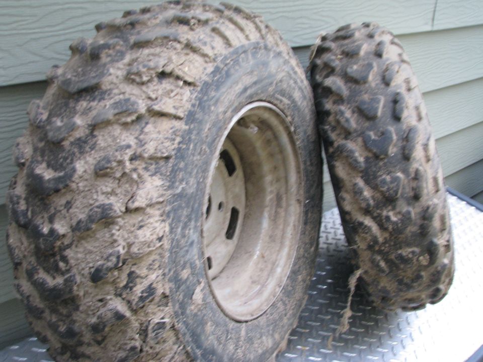 Two muddy ATV knobby tires on white steel wheels leaning against siding