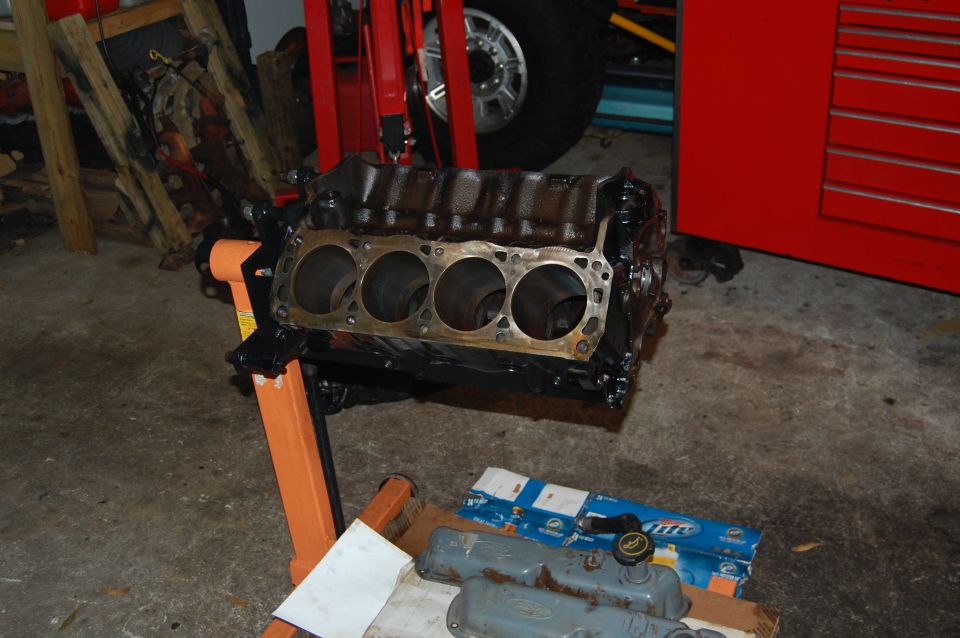 Engine block on an orange engine stand, with valve covers and a box of parts on the floor