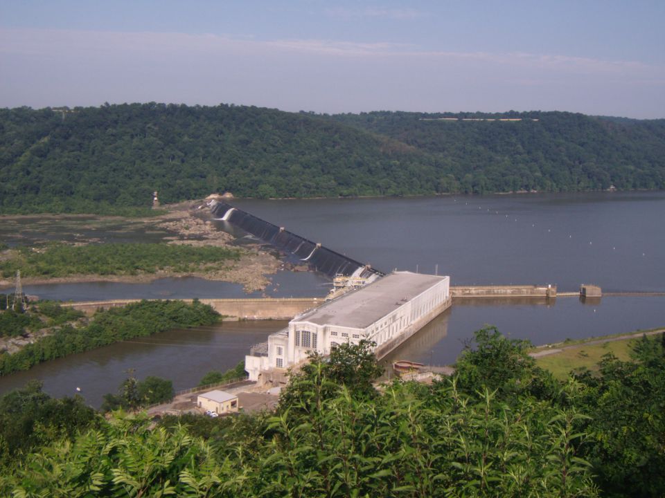 Hydroelectric dam with a long spillway and a large building next to the water, surrounded by trees