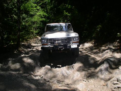 1979 Ford Bronco - A white Ford Bronco navigates a rocky, off-road trail surrounded by dense forest.