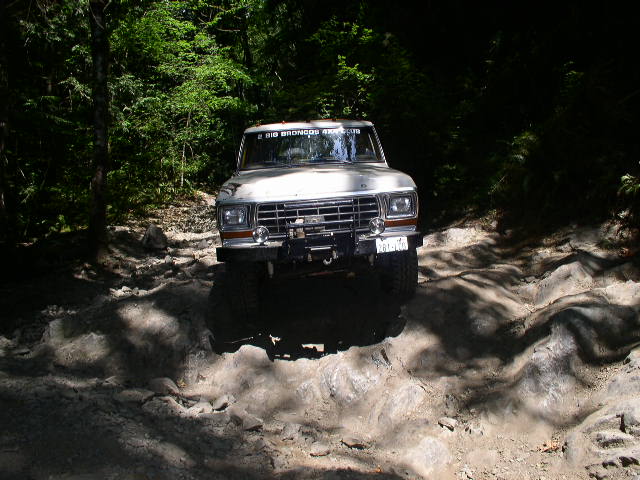 White Ford Bronco driving over rocks on a wooded trail. Text on windshield reads "BIG BRONCOS 4X4"
