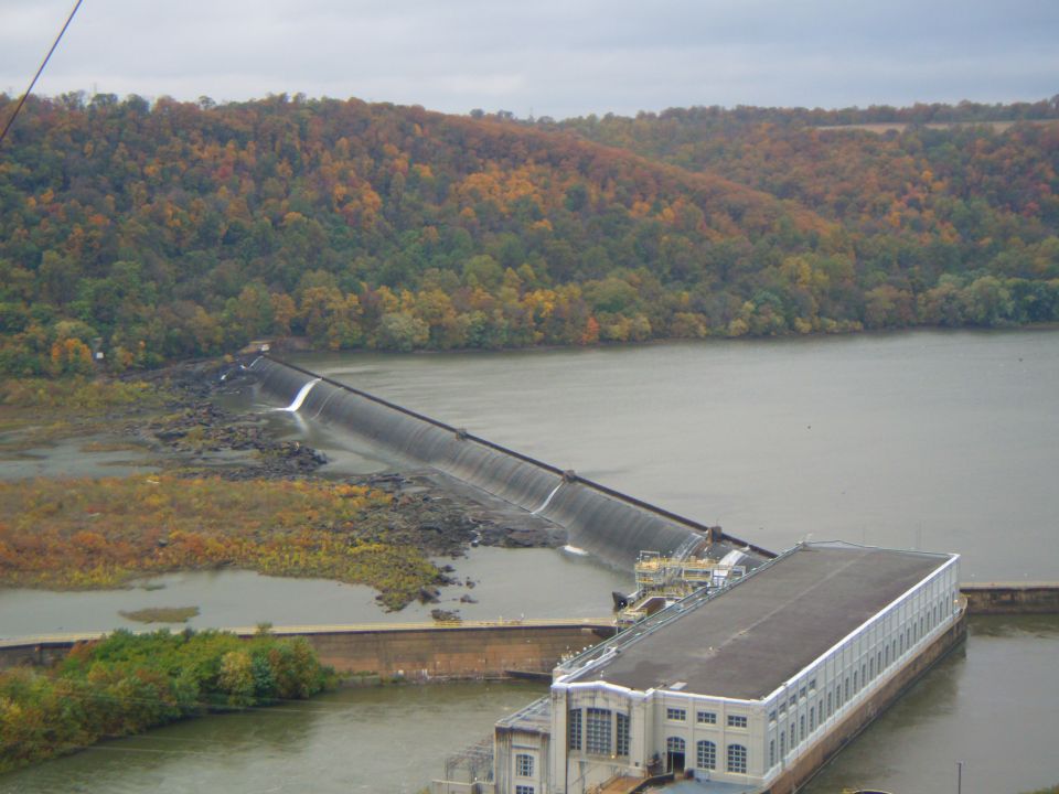 dam with water flowing over it, power station building, and autumn trees on a hill