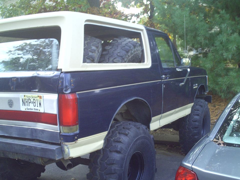 Dark blue Ford Bronco with white top, lifted with large tires. Two spare tires are visible inside the rear cargo area. New Jersey license plate NHP-81W