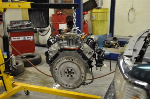 1996 Ford Bronco - A V8 engine is suspended by a chain hoist in a workshop environment, with a vehicle partially visible to the right.