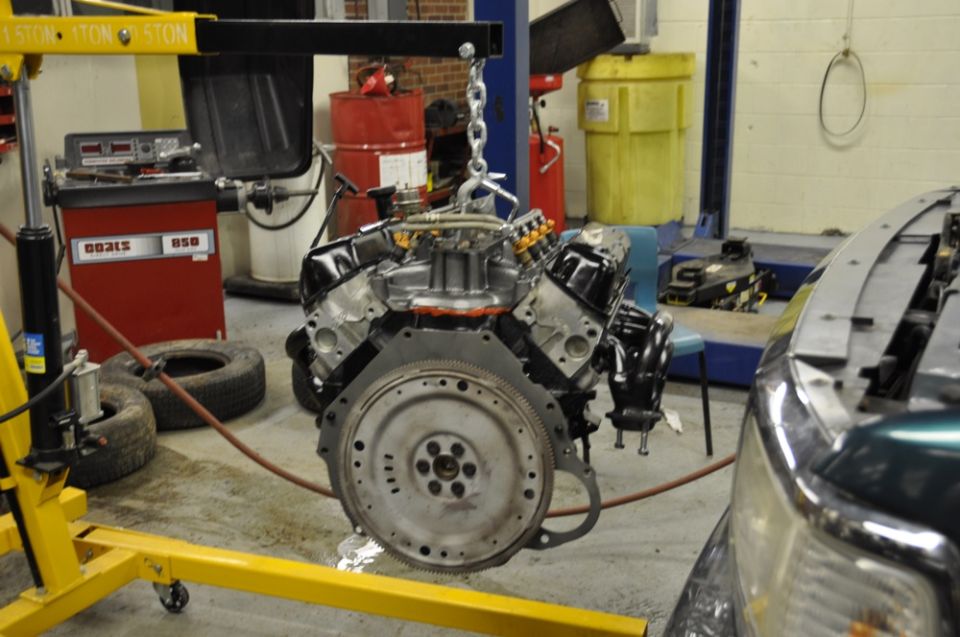 V8 engine hanging from a hoist in a garage, next to a car with its hood open
