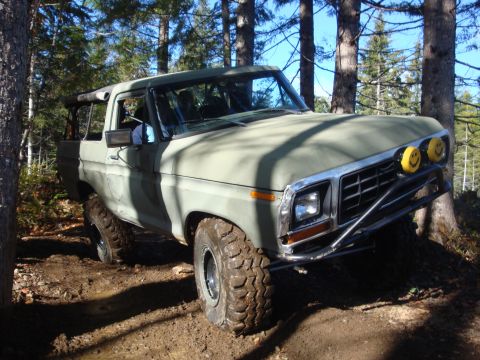 1978 Ford Bronco - Olive green 1978 Ford Bronco navigates a muddy forest trail with large off-road tires and custom smiley face headlights.