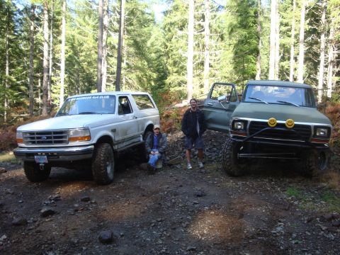 1978 Ford Bronco - Two off-road vehicles are parked on a dirt path surrounded by trees, with two people standing nearby.
