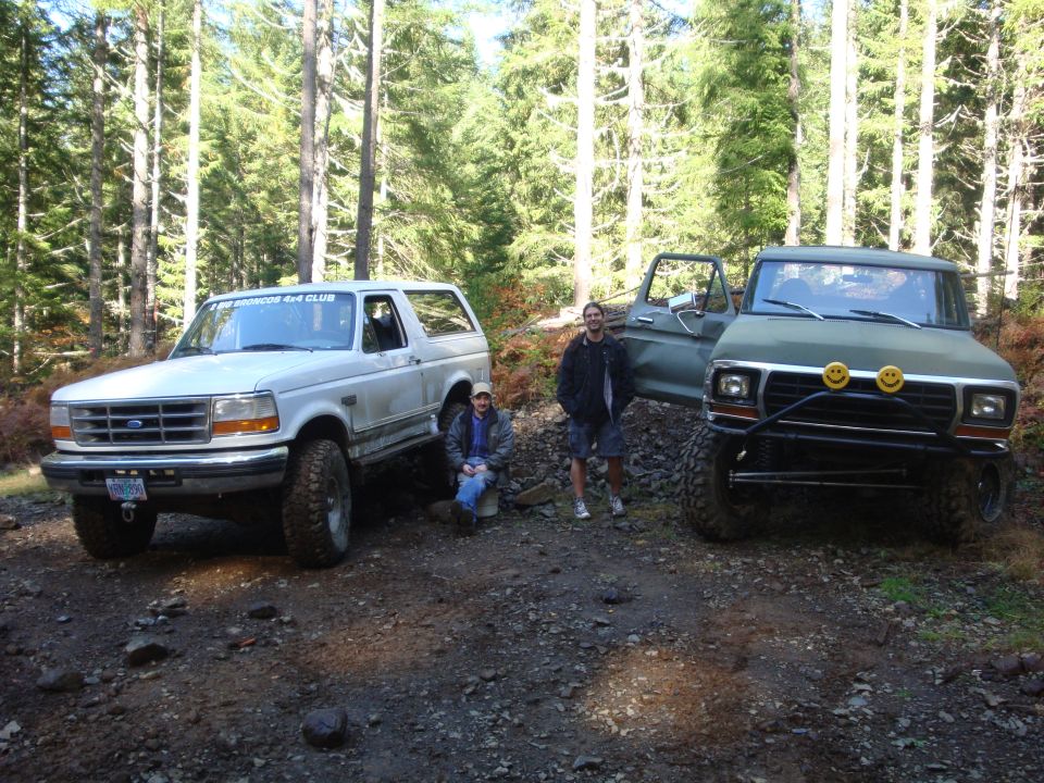 Two people with a white Bronco and a green truck in a wooded area