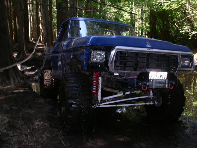 Blue off-road SUV with large tires and winch, angled from the front-right in a muddy forest