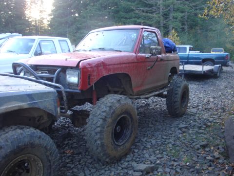 1978 Ford Bronco - Modified red pickup truck with off-road tires and a brush guard parked on gravel.