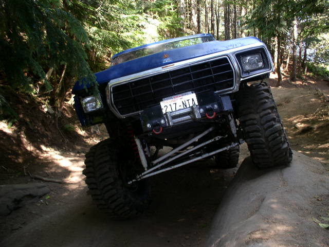 Blue 1979 Ford Bronco climbing over a large rock on a dirt trail, trees in background