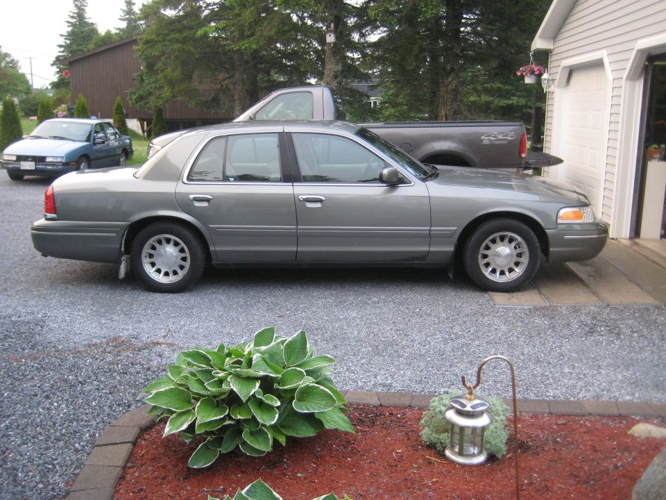 Side view of gray-green Crown Victoria sedan on gravel driveway next to garage, truck and car in background