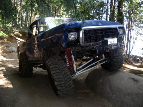 1979 Ford Bronco - A heavily modified blue Ford Bronco navigates a challenging off-road course with its tires at extreme angles.
