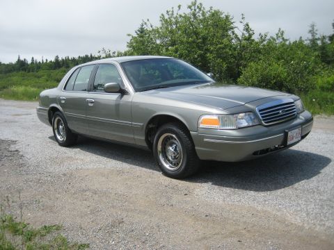 1999 Ford Crown Victoria - A gray sedan sits on a gravel lot with trees in the background.