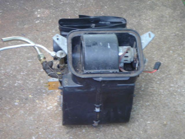 Removed black heater box assembly with blower motor and heater core tubes on concrete