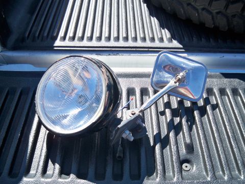 1985 Ford Bronco - A detached headlight and side mirror are placed on the textured surface of a truck bed.