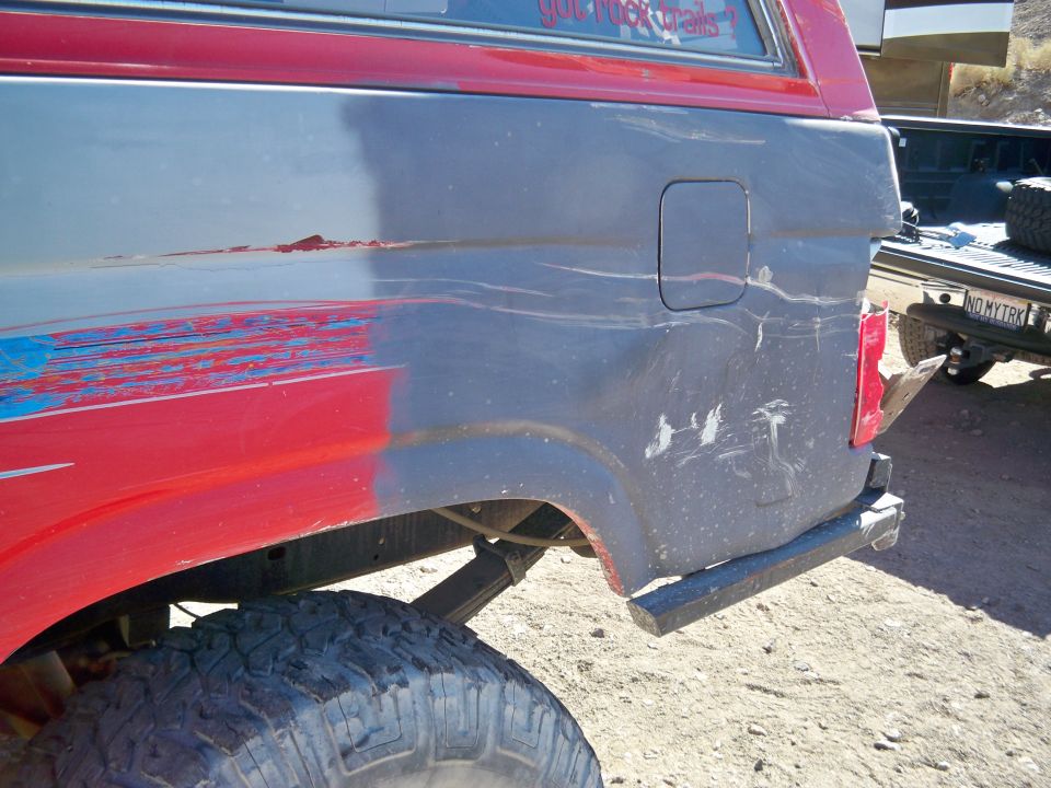Rear quarter panel of a red and gray SUV with visible damage and a fuel door. Another vehicle is partially visible in the background