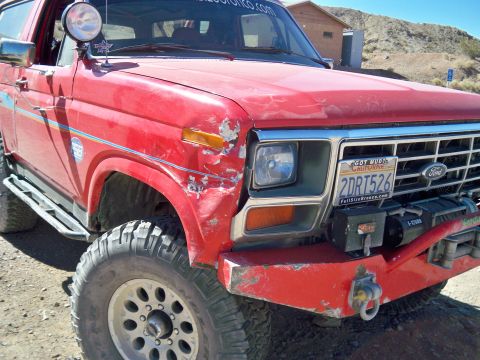 1985 Ford Bronco - This red 1985 Ford Bronco features a dented front fender, off-road tires, and a winch, suggesting it's built for terrain.