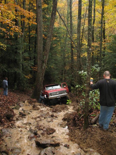 1985 Ford Bronco - A red pickup truck is stuck in a muddy, rocky trail surrounded by trees with autumn foliage.
