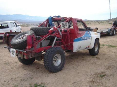 1984 Ford Bronco - Modified off-road truck with extensive roll cage and spare tires is parked in a desert landscape next to another vehicle and trailer.