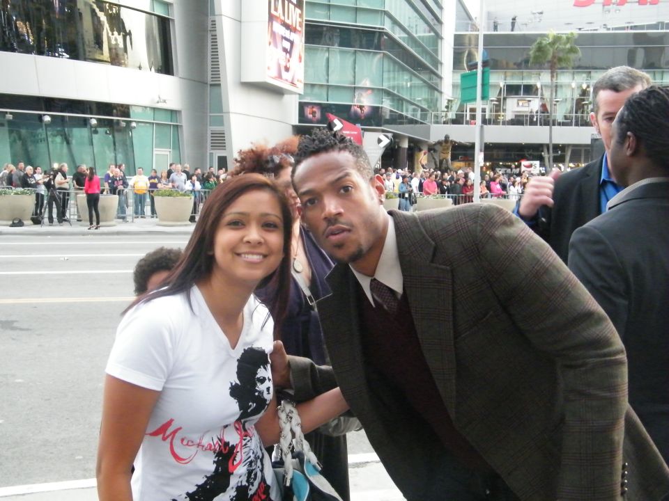 Woman in white t-shirt and man in suit pose for photo outside building with crowd in background