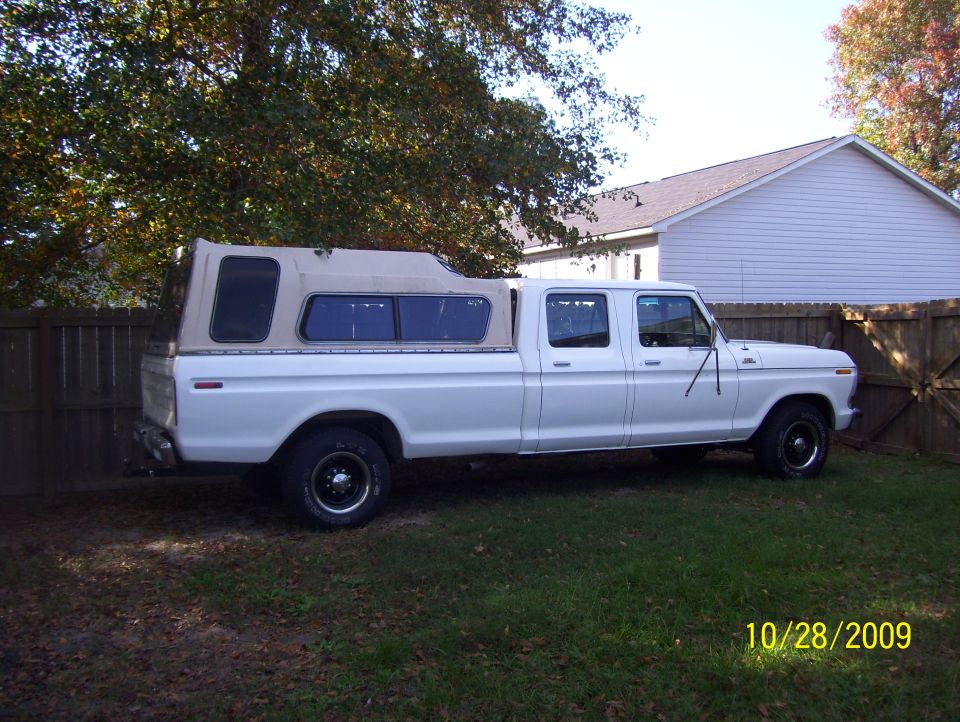 Side profile of white 1970s Ford crew cab long-bed pickup with camper shell in yard