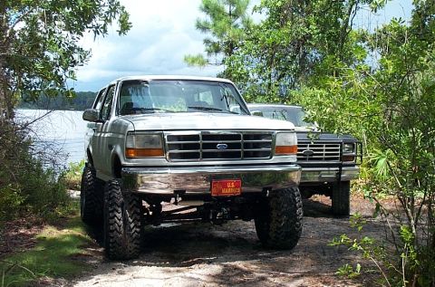 1995 Ford Bronco - Two Ford Broncos are parked off-road near a lake, one is lifted with custom wheels.