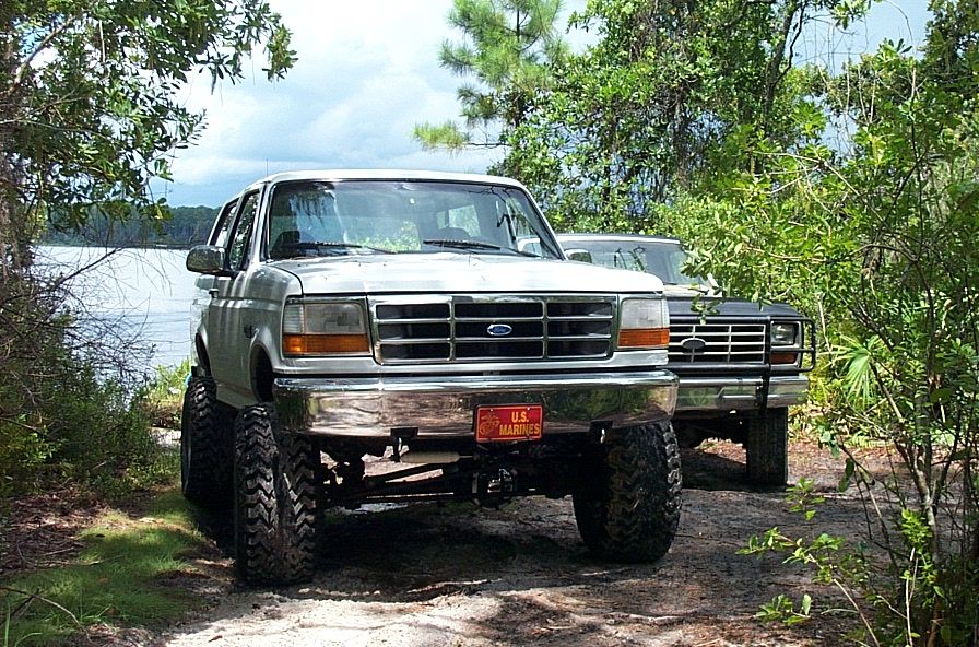Lifted white Ford Bronco with U.S. Marines front plate parked on a sandy trail beside a lake