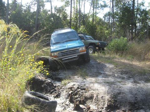 1992 Ford Explorer - A teal 1992 Ford Explorer navigates a challenging muddy path, with another SUV visible in the background.