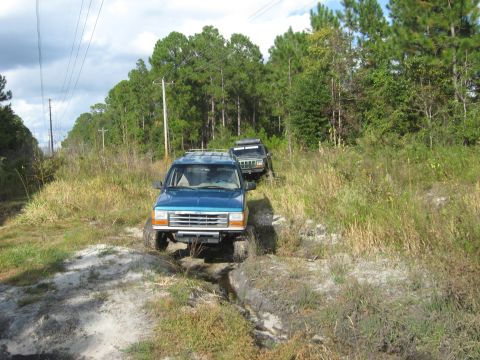 1992 Ford Explorer - Two SUVs navigate a rough dirt trail surrounded by trees and tall grass.