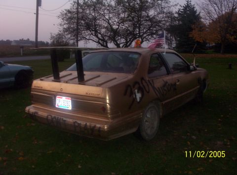 1982 Oldsmobile Cutlass - A gold sedan with a large rear spoiler and graffiti is parked outdoors on grass.