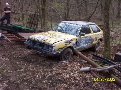 1982 Oldsmobile Cutlass - A yellow car with graffiti is parked in a wooded area next to a trailer.