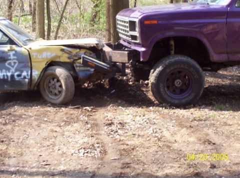 1982 Oldsmobile Cutlass - A yellow car with graffiti and a damaged front end is being pushed by a purple lifted truck in a wooded area.