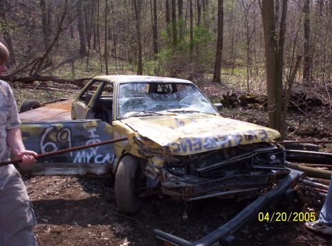 1982 Oldsmobile Cutlass - A heavily damaged yellow car sits in a wooded area, showing graffiti on its doors and hood, a broken windshield.