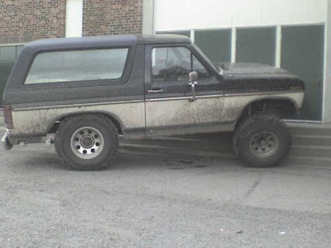1985 Ford Bronco - A dirty, two-tone Ford Bronco with a camper shell is parked on a gravel surface.