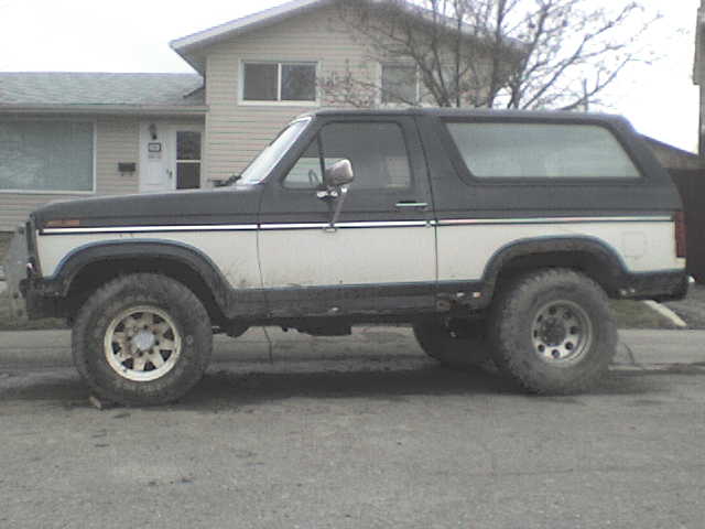 Side view of a two-tone Ford Bronco with a black top and white body. It has large off-road tires and is parked in front of a house
