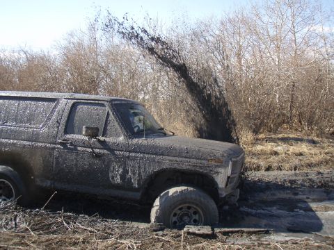 1985 Ford Bronco - A heavily mud-caked Ford Bronco is stuck in a muddy area with trees in the background.