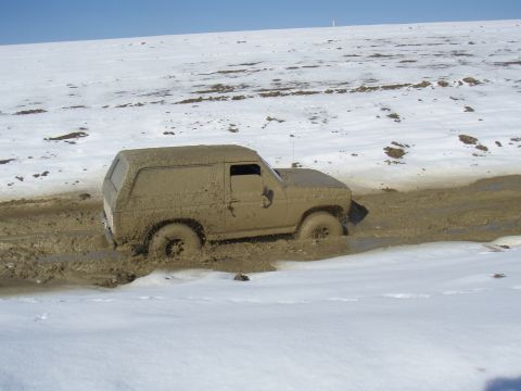 1985 Ford Bronco - This 1985 Ford Bronco is deeply stuck in a muddy terrain, with snow visible on the surrounding hills.