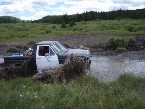 1985 Ford Bronco - A pickup truck navigates a muddy waterway, kicking up a large spray.