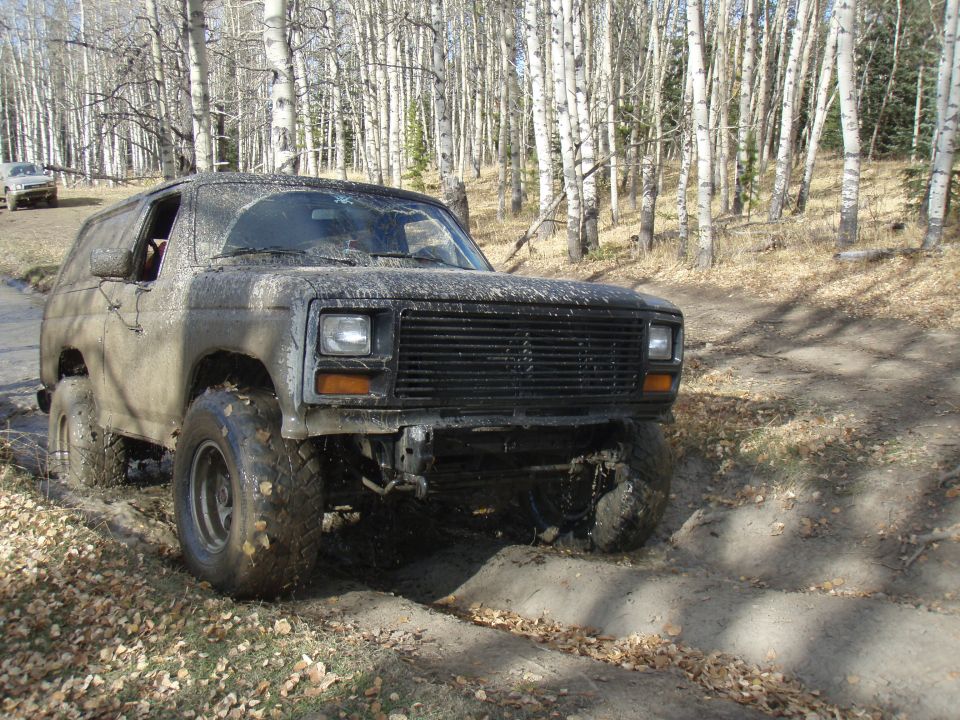Mud-covered SUV with a black grille and square headlights, driving on a dirt path in a forest. Another vehicle is visible in the background