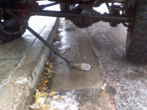 1993 Ford Bronco - A vehicle's undercarriage is visible, with a jack positioned near a wet curb and fallen leaves.