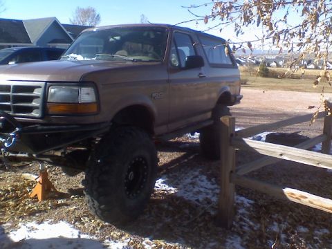 1993 Ford Bronco - Brown 1993 Ford Bronco with aftermarket bumper and large tires, parked on uneven ground.