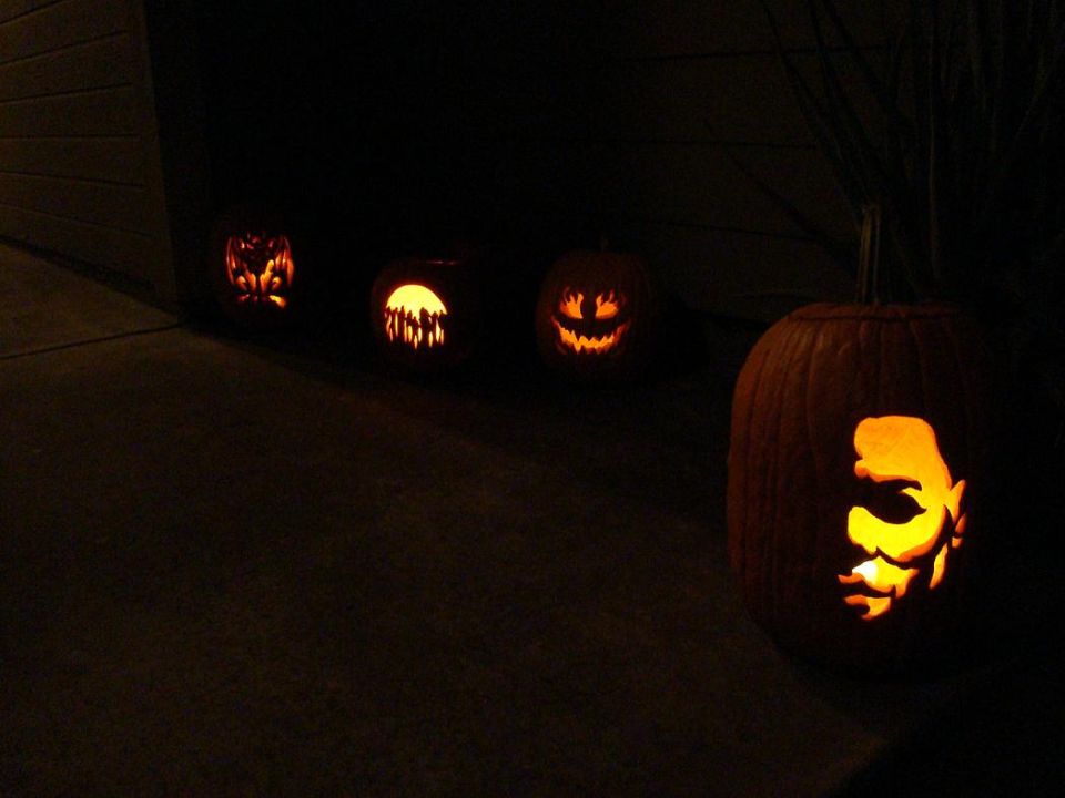 Four carved pumpkins lit up at night. One pumpkin in the foreground is carved with a face. Three other pumpkins are in the background
