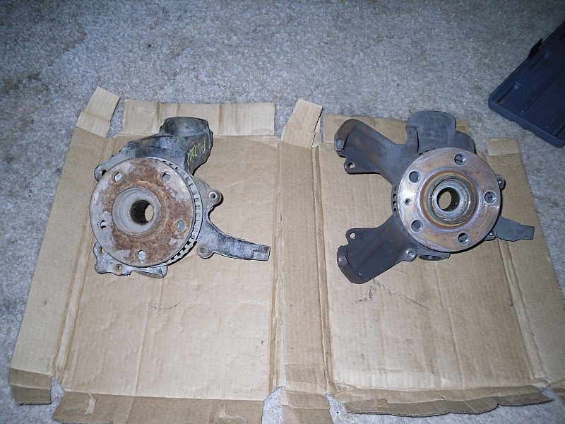 Two automotive steering knuckles with brake rotors, one rusty, laid on cardboard