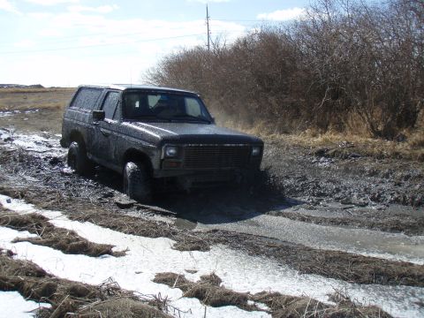 1985 Ford Bronco - A dark SUV navigates a challenging muddy terrain with patches of ice and snow.